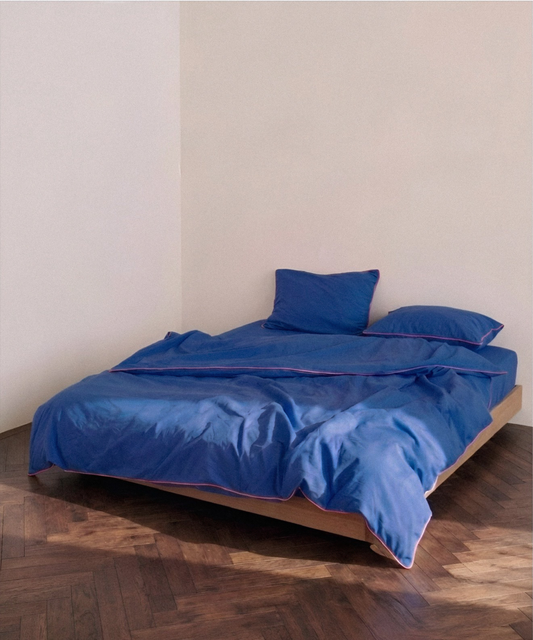 A bed dressed in a royal blue bedding set with matching pillows, set on a wooden platform bed frame in a minimalist room with beige walls and a dark wood parquet floor.