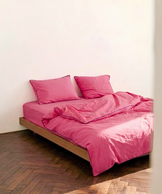 A bed dressed in a bright pink bedding set with matching pillows and duvet, set on a wooden platform bed frame against a plain off-white wall with a dark wood parquet floor.