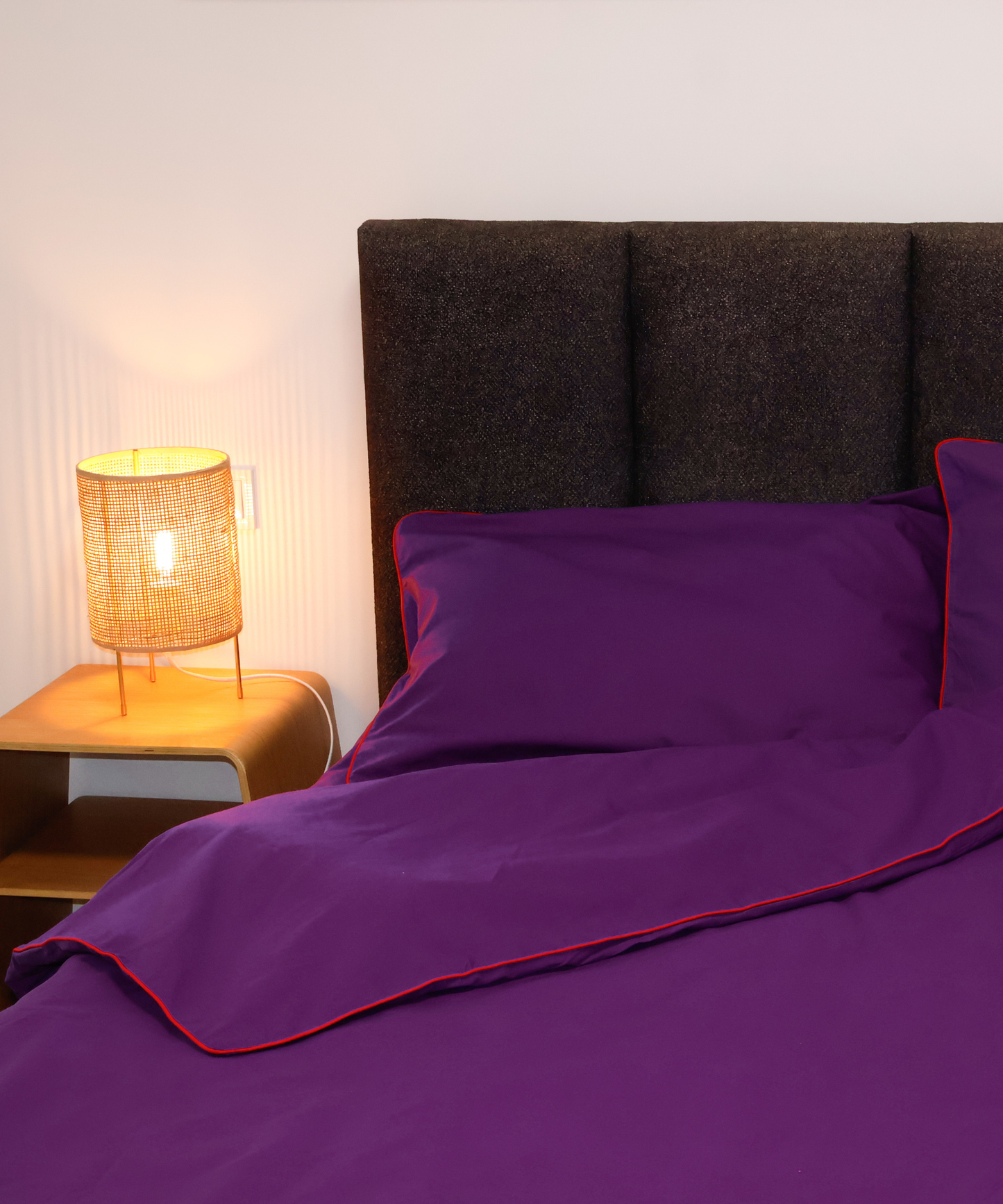Purple bedding set with red piping trim, styled on a bed with a dark textured headboard and a lit bedside lamp.