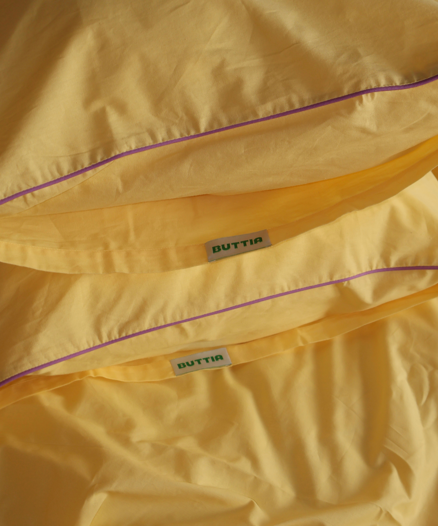 Close-up of yellow bedding, likely a duvet cover or comforter, with a purple piping accent and two visible Buttia brand labels.