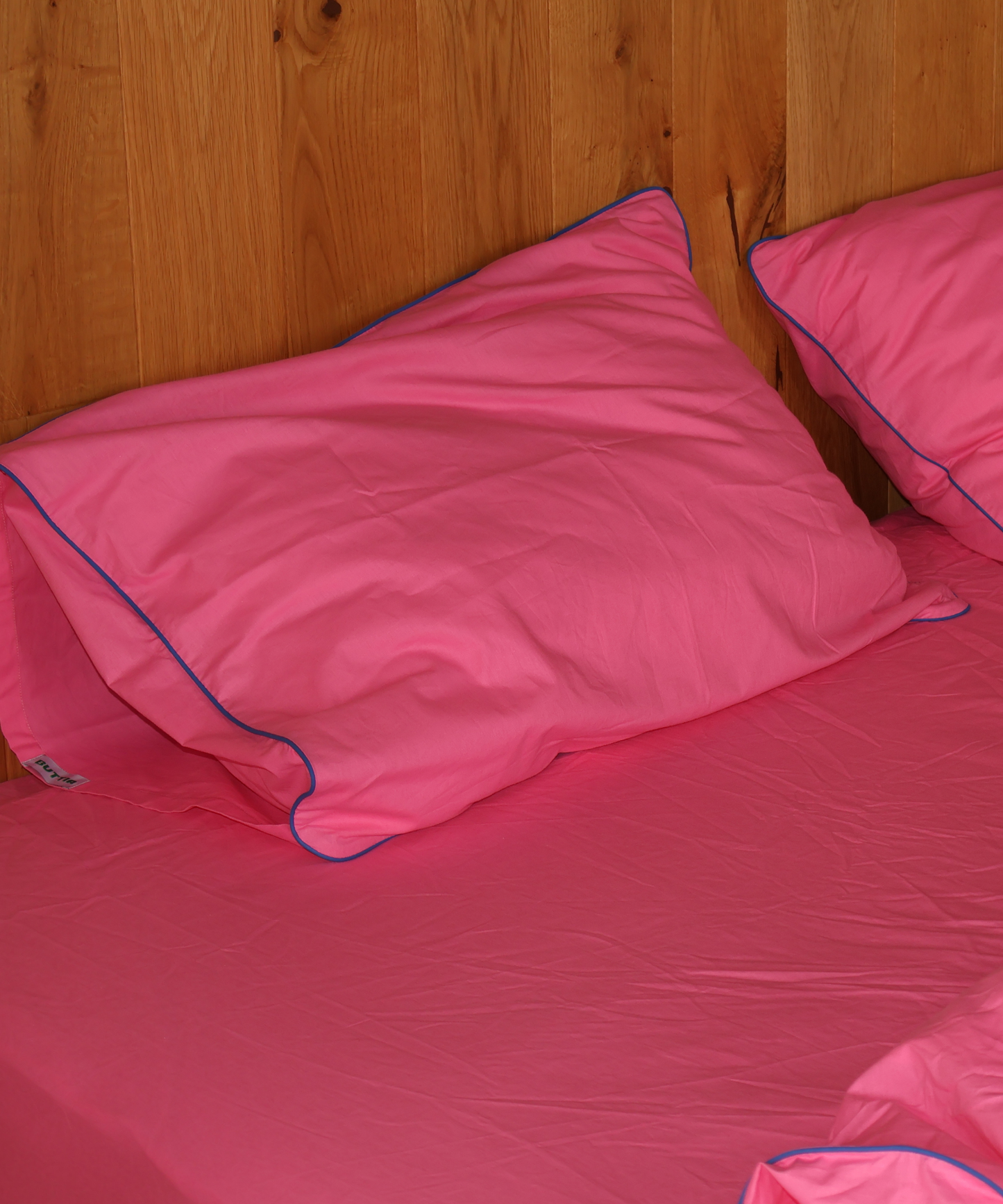 Bright pink bedding set with matching pillowcases and a fitted sheet, featuring contrasting blue piping trim, shown against a wooden headboard.