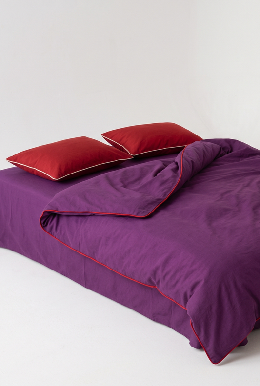 Purple bedding set with a duvet cover and two red pillows, featuring red piping trim, shown on a bed against a plain light gray background.
