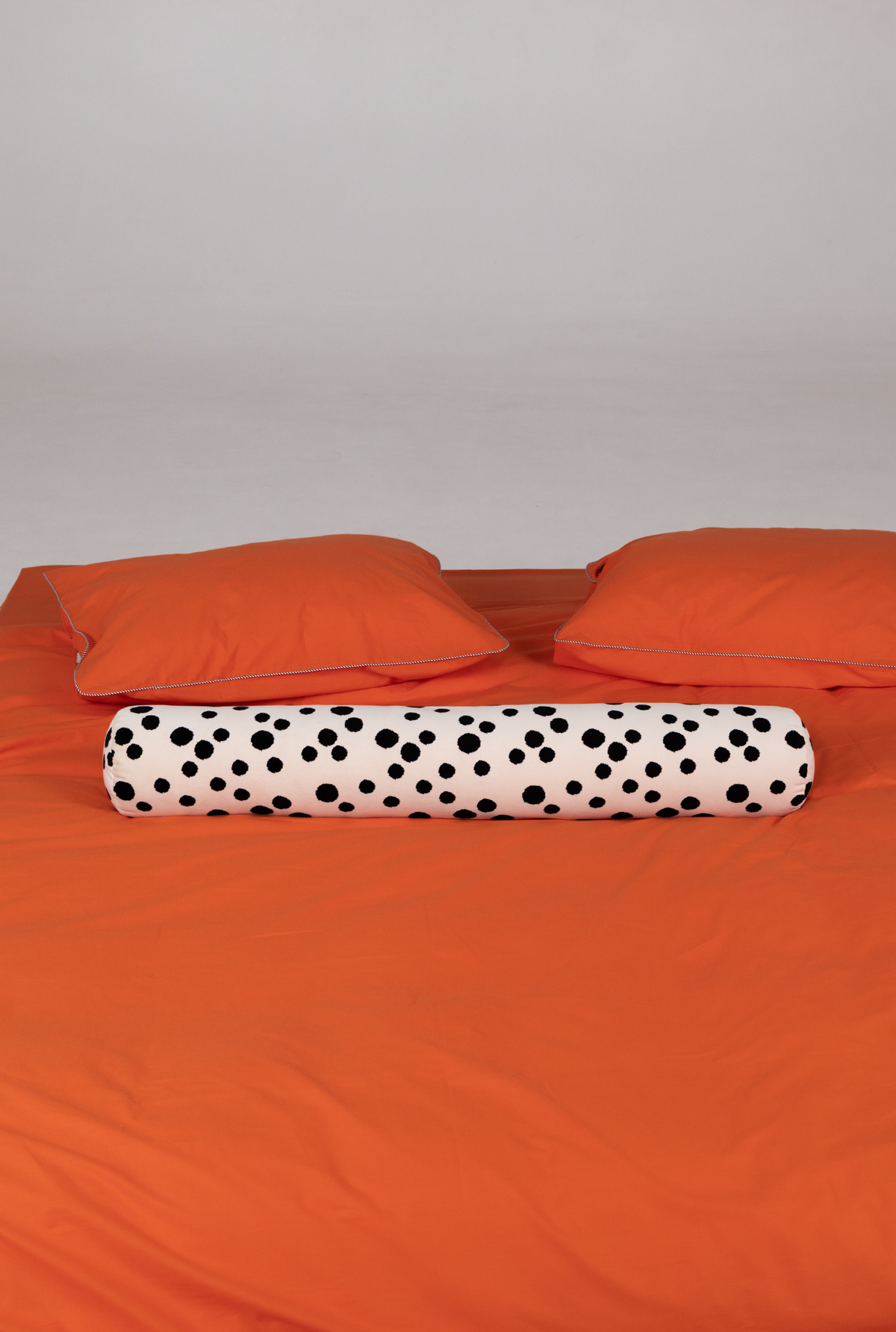 An orange bedding set on a bed, featuring an orange duvet cover, two matching orange pillows, and a cylindrical lumbar pillow with a black polka dot pattern on a white background.