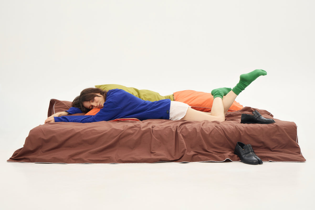 A person lies on a bed with a brown sheet BUTTIA, wearing a blue top, orange pants, white shorts, green socks, and black shoes, photographed against a plain white studio background.