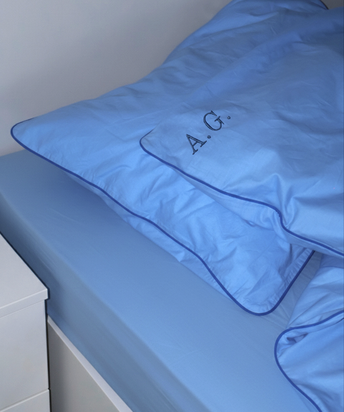A light blue bedding set on a bed, featuring a pillowcase with dark blue piping and the monogram 'A.G.