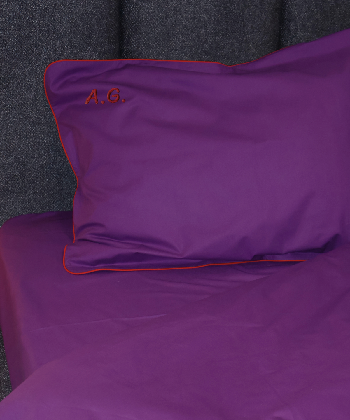 A close-up of a purple pillowcase and matching bed sheet set with red piping trim and an embroidered 'A.G.