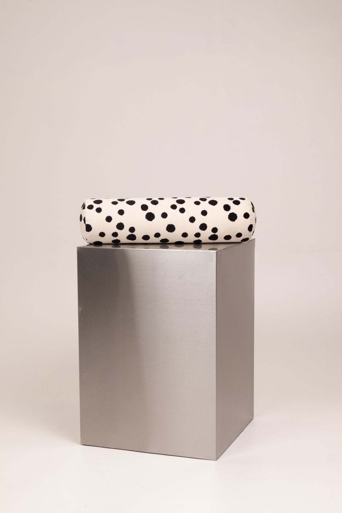 A folded blanket with a black dalmatian spot pattern on a white background, displayed on a metallic silver cube pedestal against a light gray studio background.