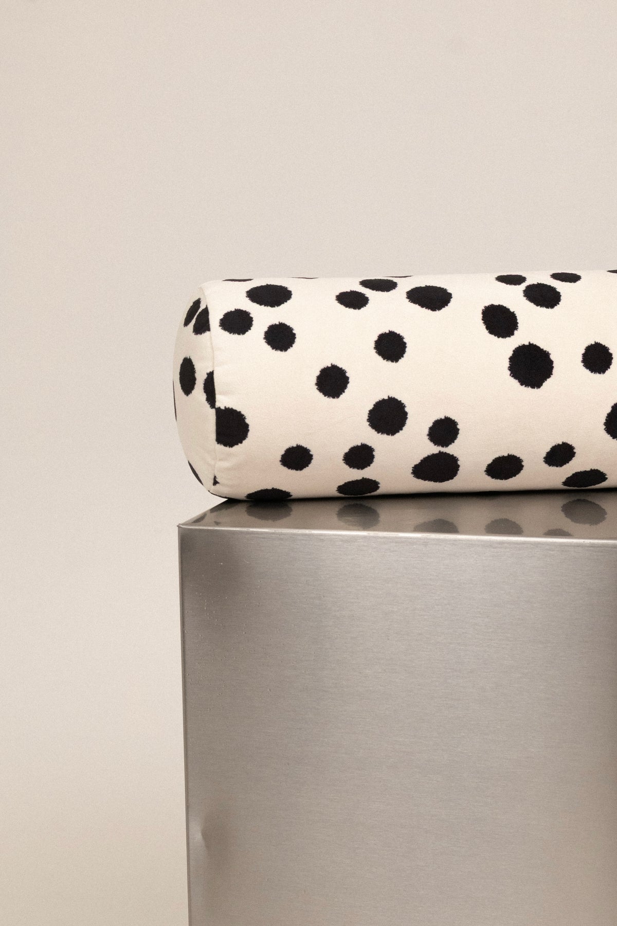 A cylindrical lumbar pillow with a black dalmatian spot pattern on a cream background, resting on a metallic silver pedestal against a light gray wall.