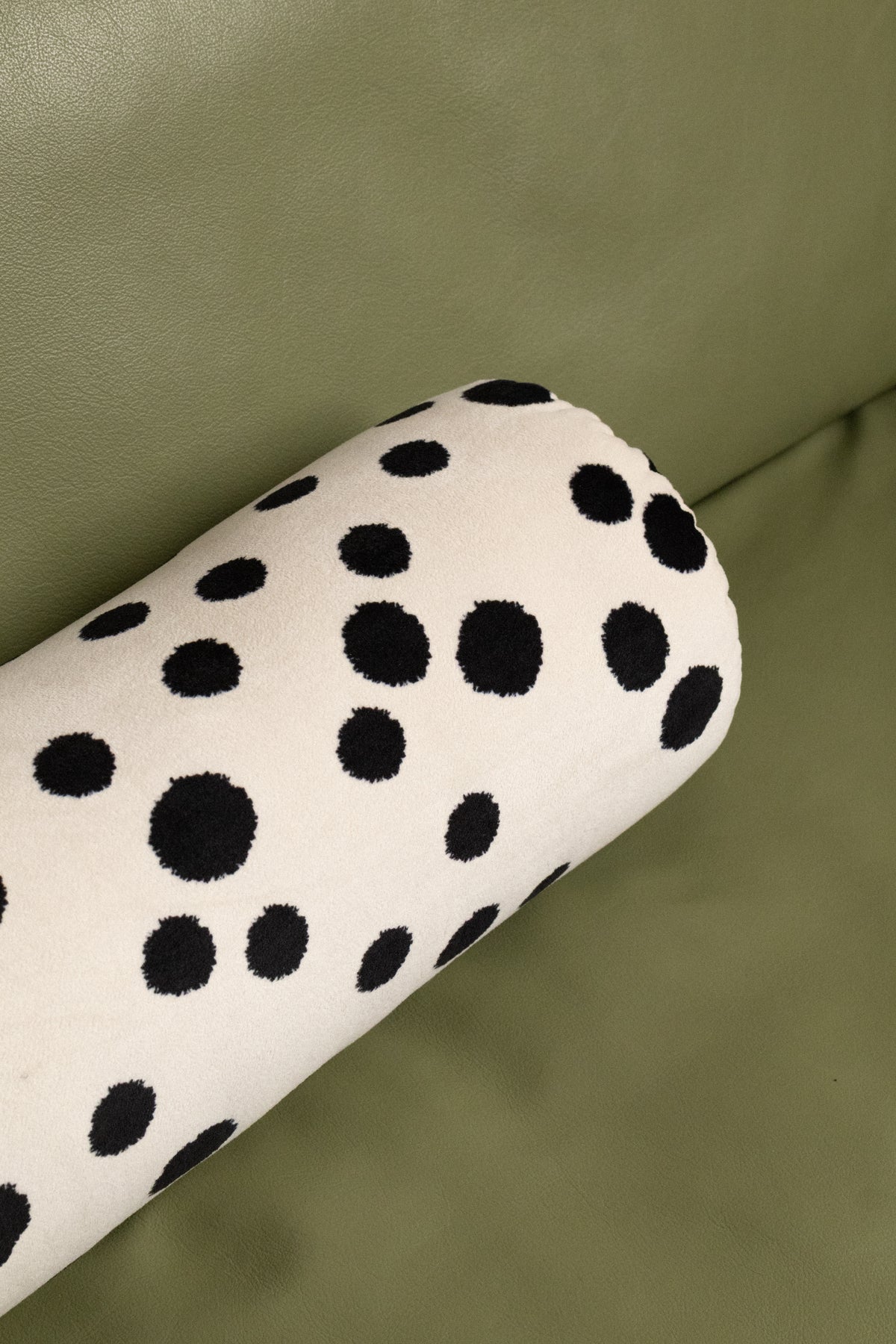 A close-up of a cylindrical cushion or bolster with a black dalmatian spot pattern on a cream fabric, resting on an olive green leather surface.
