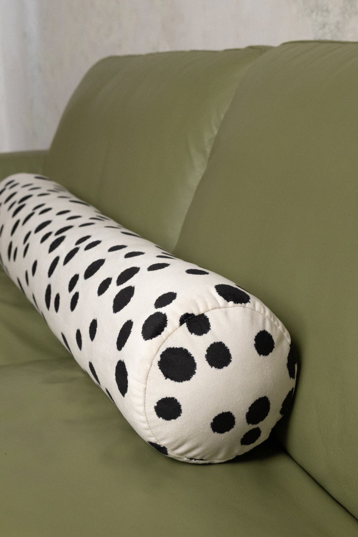 A cylindrical lumbar pillow with a black and white polka dot pattern, resting on an olive green sofa.