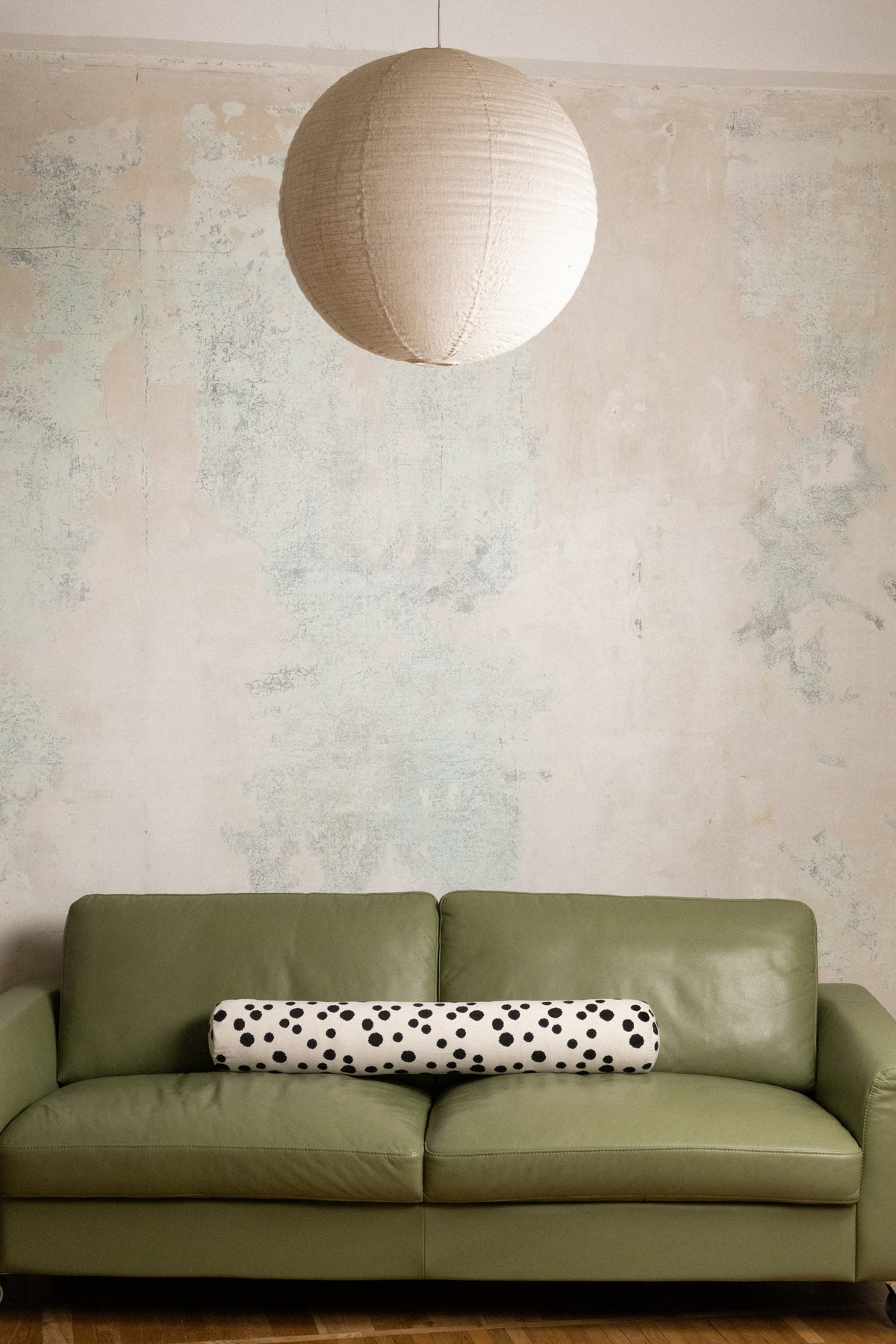 An olive green leather sofa in a styled room setting, featuring a black and white polka dot cylindrical lumbar pillow and a large spherical paper lantern hanging from a textured plaster wall.