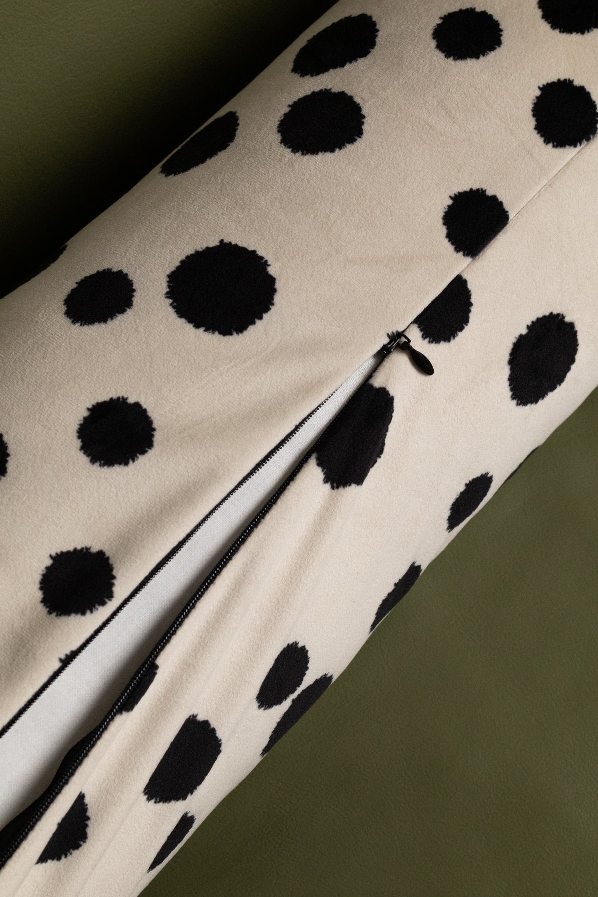 Close-up of a cream-colored fabric item with a black dalmatian-style spot pattern and a visible zipper, photographed against an olive green background.