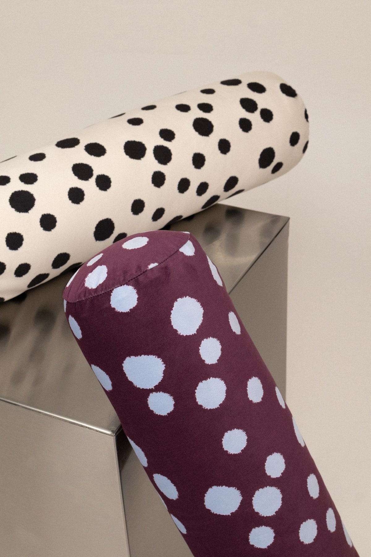 Two cylindrical bolster pillows with a polka dot pattern, one in cream with black dots and the other in plum with light blue dots, displayed on a metallic cube against a neutral background.