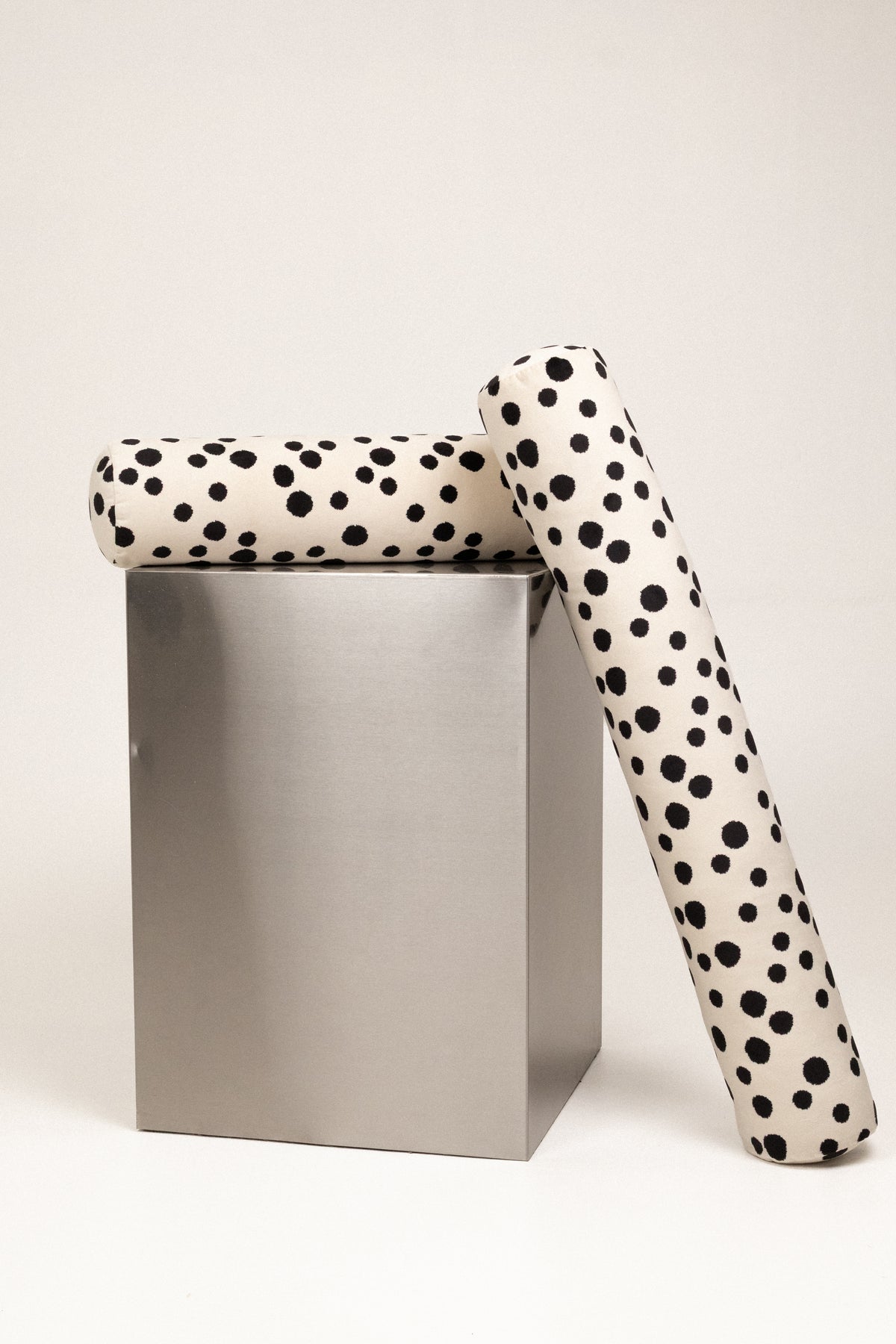 Two rolled pieces of fabric or upholstery with a black-on-white dalmatian spot pattern, displayed on and beside a metallic silver cube against a light neutral background.