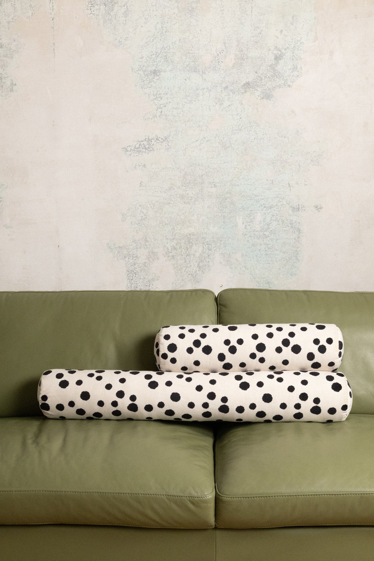 Two cylindrical lumbar pillows with a black and white dalmatian spot pattern, stacked on an olive green leather sofa against a textured light gray wall.