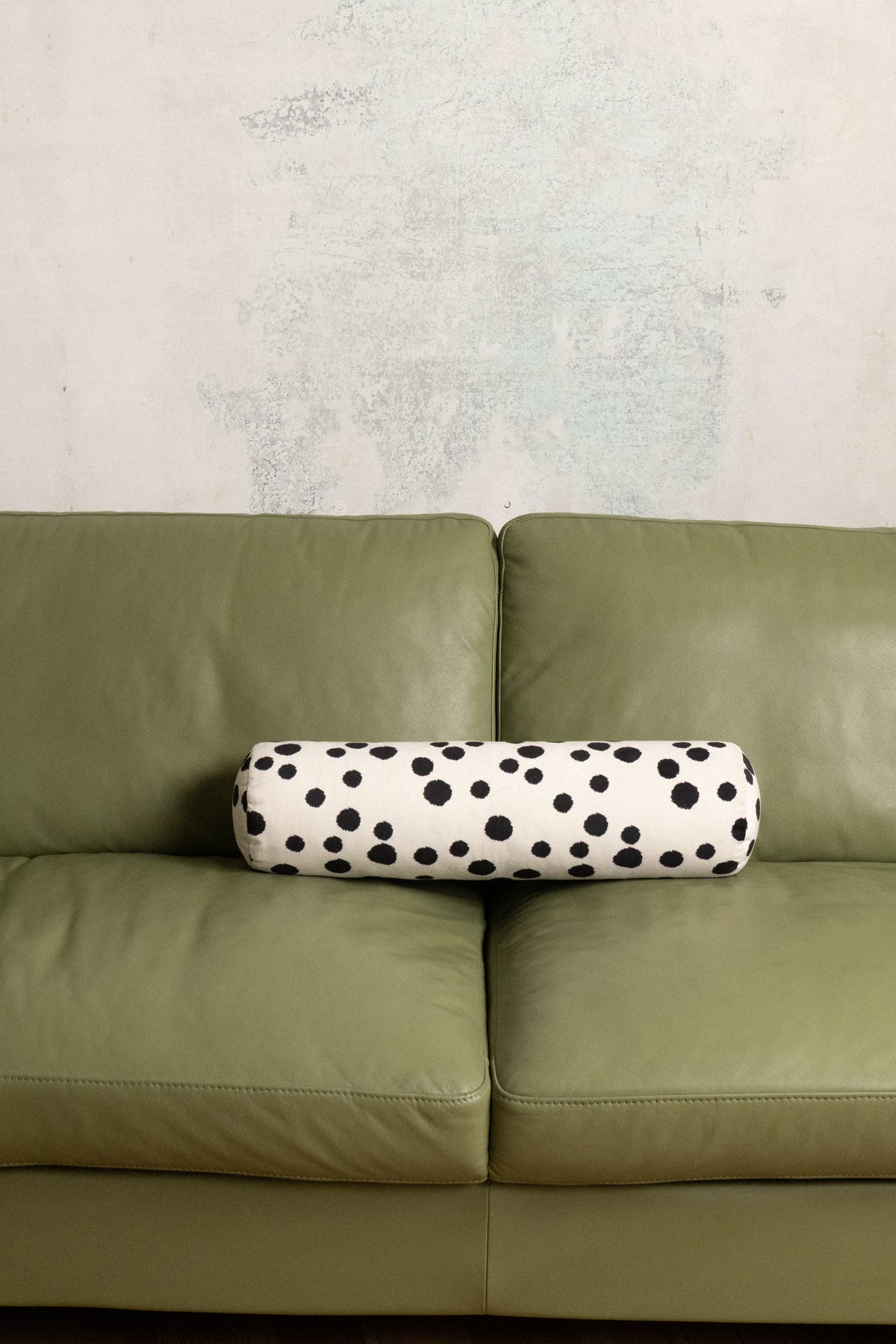 A cylindrical lumbar pillow with a black and white dalmatian spot pattern, resting on the seat of an olive green leather sofa against a textured light gray wall.