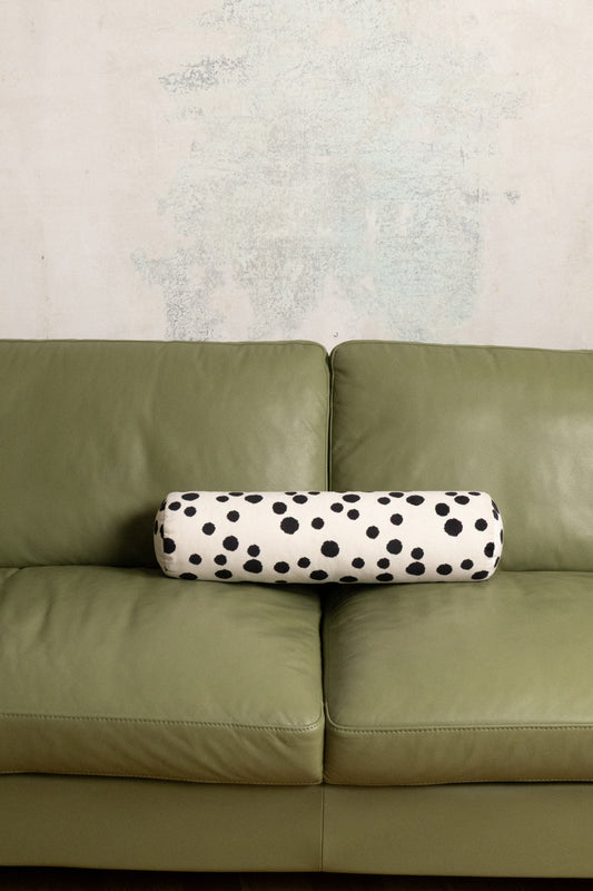 A cylindrical lumbar pillow with a black and white dalmatian spot pattern, resting on the seat of an olive green leather sofa against a textured light gray wall.
