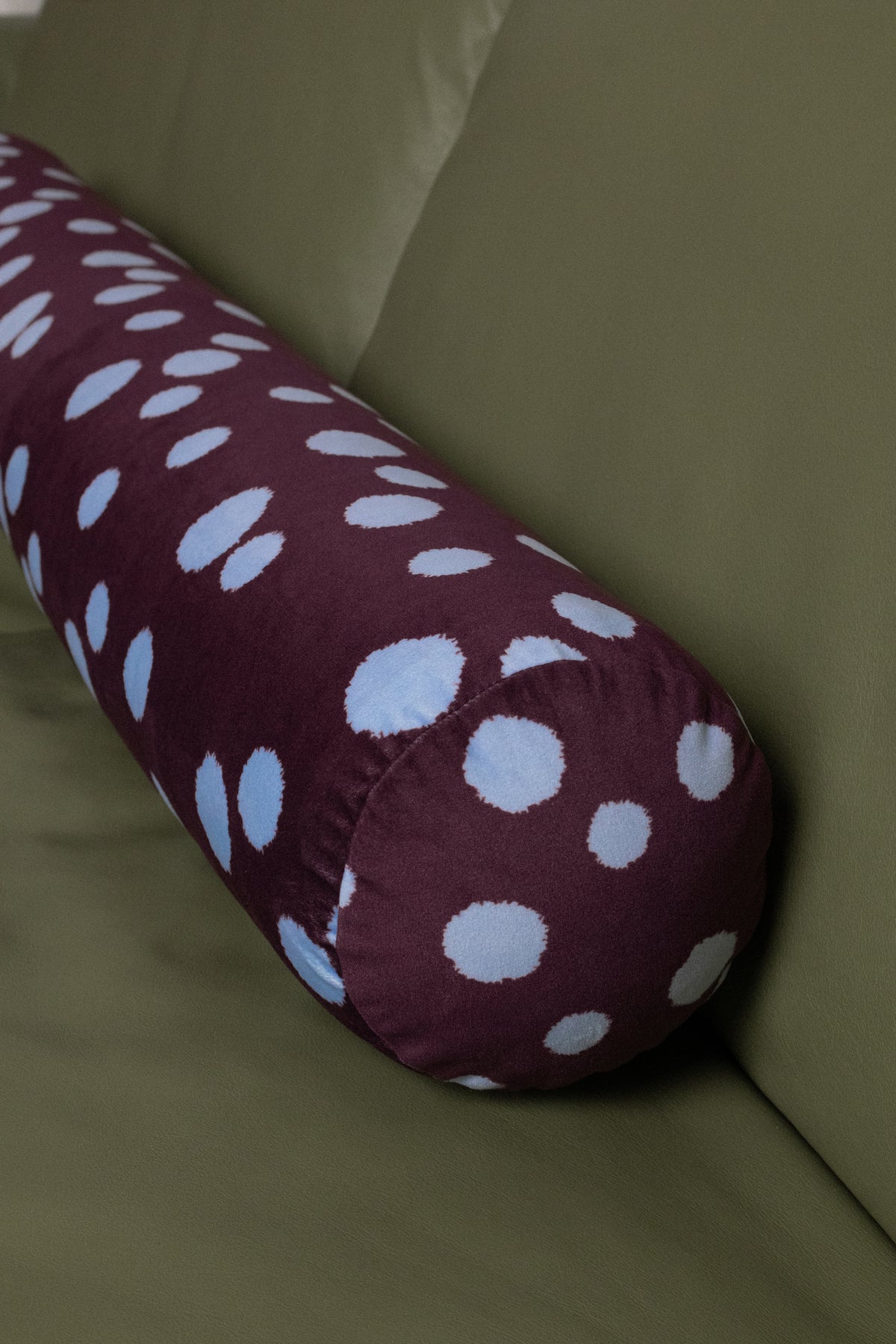 A cylindrical bolster pillow with a plum base and blue polka dots, resting on an olive green sofa.