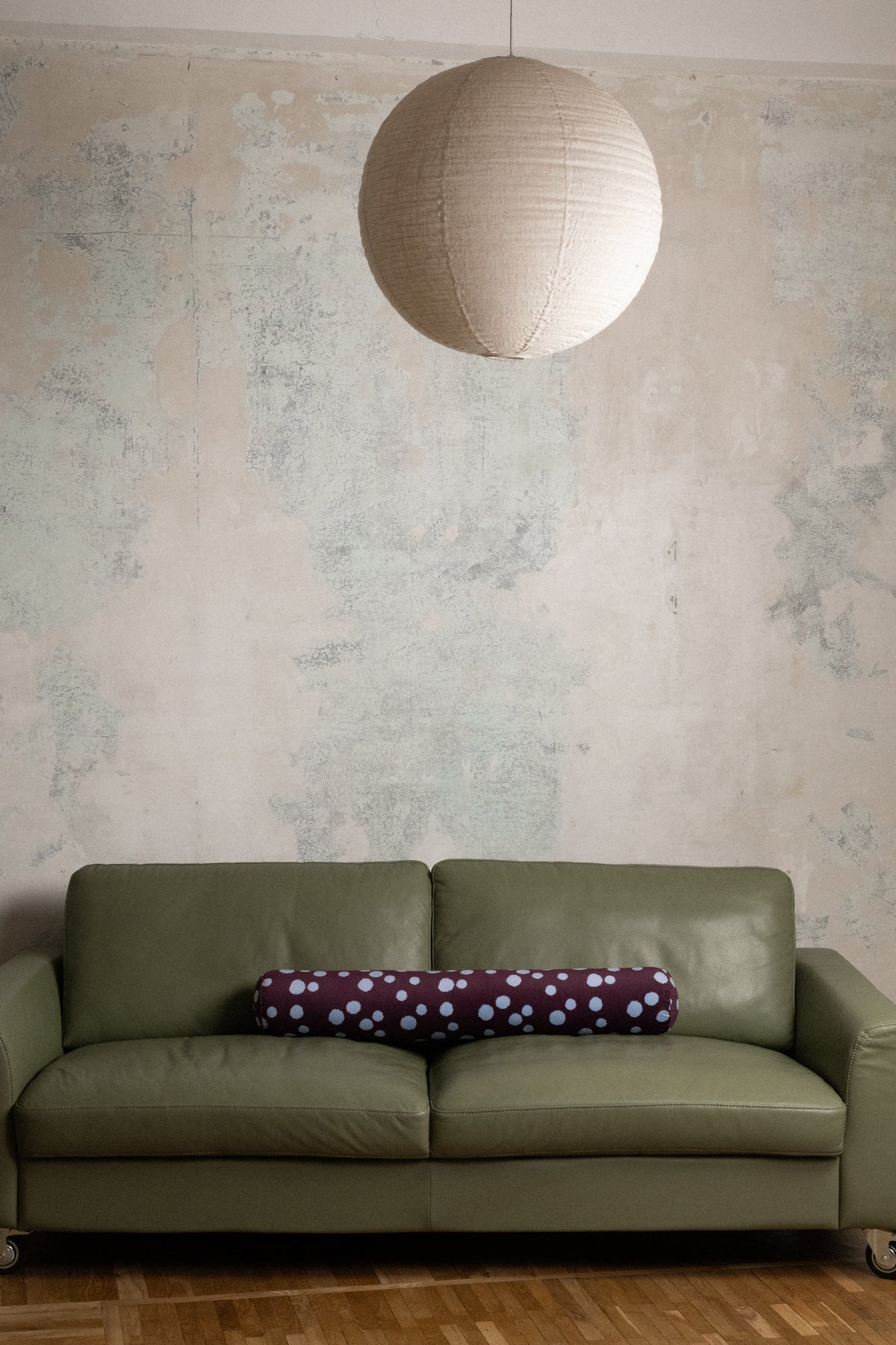 An olive green leather sofa in a rustic room setting with a textured plaster wall, a hanging paper lantern, and a purple polka-dot cylindrical pillow.