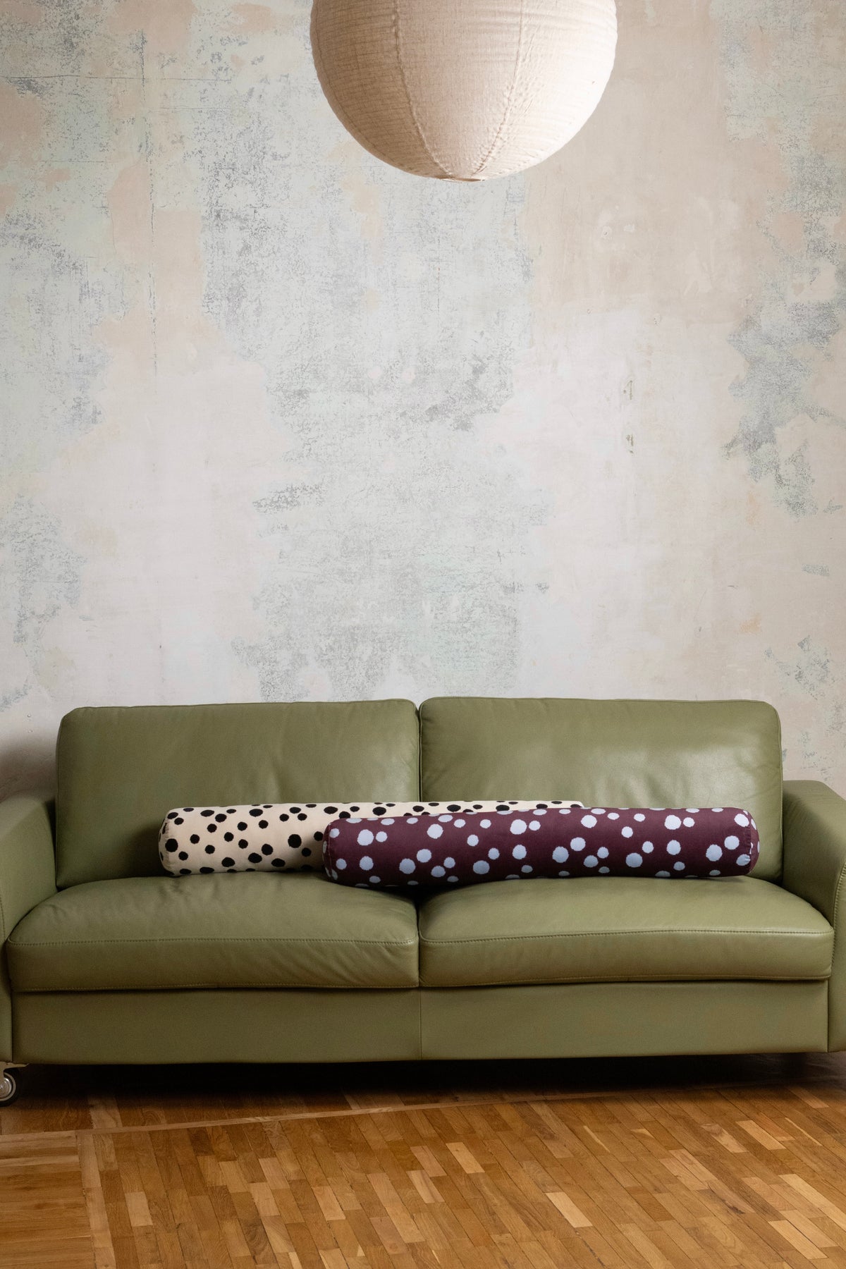 An olive green leather sofa in a styled room setting with a textured plaster wall, a hanging paper lantern, and a wood floor. Two decorative cylindrical pillows rest on the sofa: one cream with black polka dots and one plum with light blue polka dots.