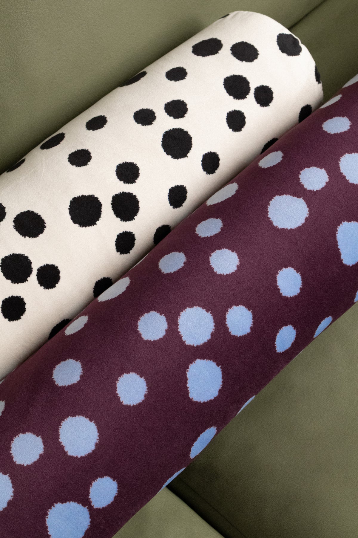 Two rolled fabric or textile samples resting on a green surface. One roll is off-white with large black polka dots, and the other is deep purple with large light blue polka dots.