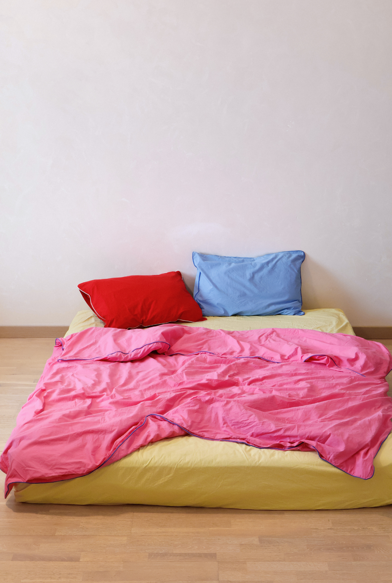 Bed with pink comforter, red and blue pillows on a wooden floor.