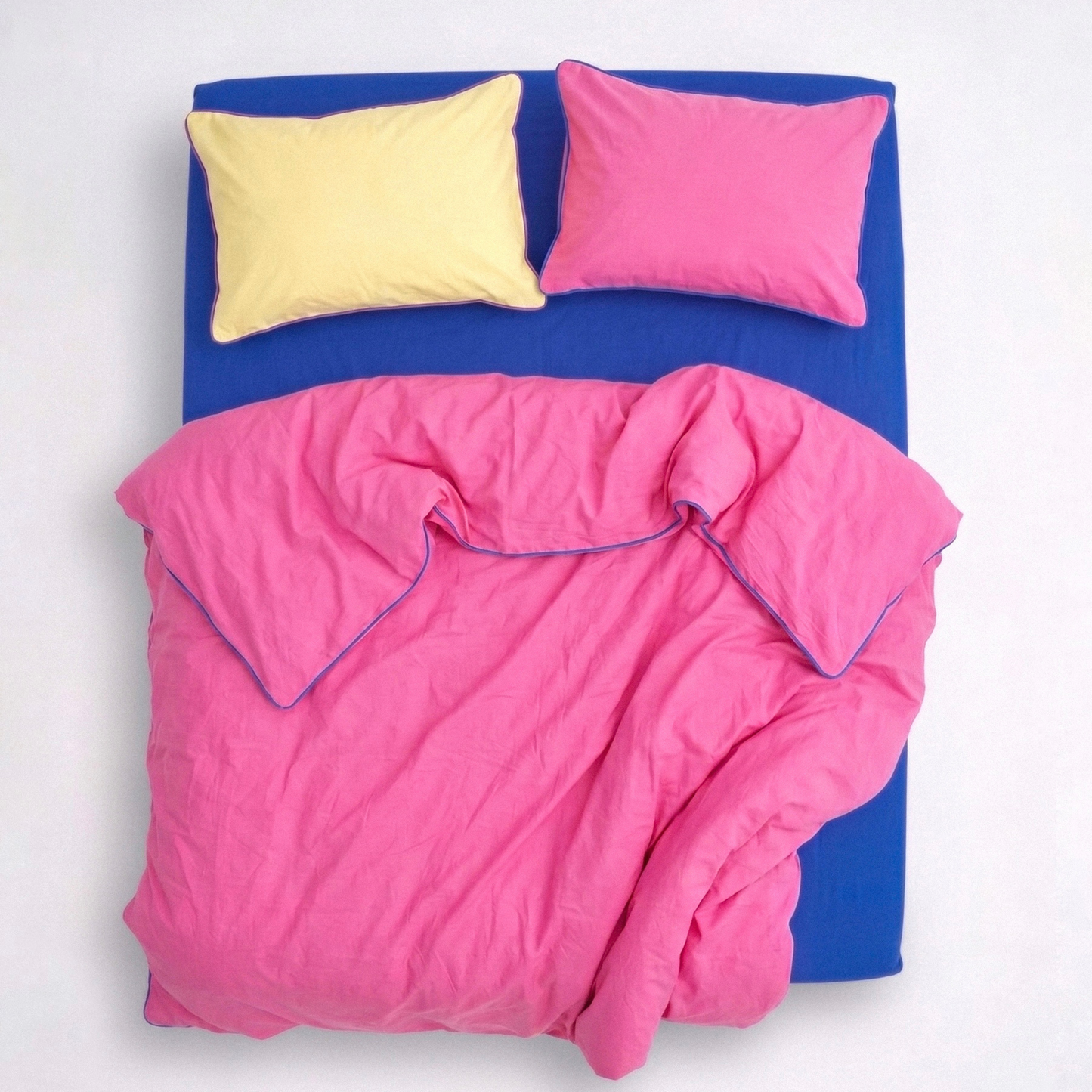 Hot pink and blue bedding set with yellow and pink pillowcases, view from above.Hot pink and blue bedding set with yellow and pink pillowcases, view from above. BORDERLINE Palette 05 by BUTTIA, handmade cotton bedding with accent piping.