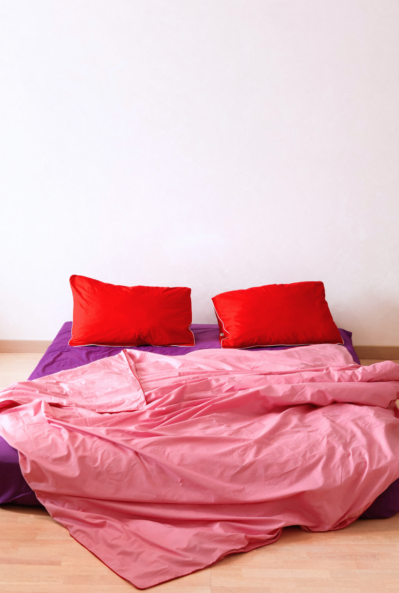 Bed with pink comforter and red pillows against a white wall