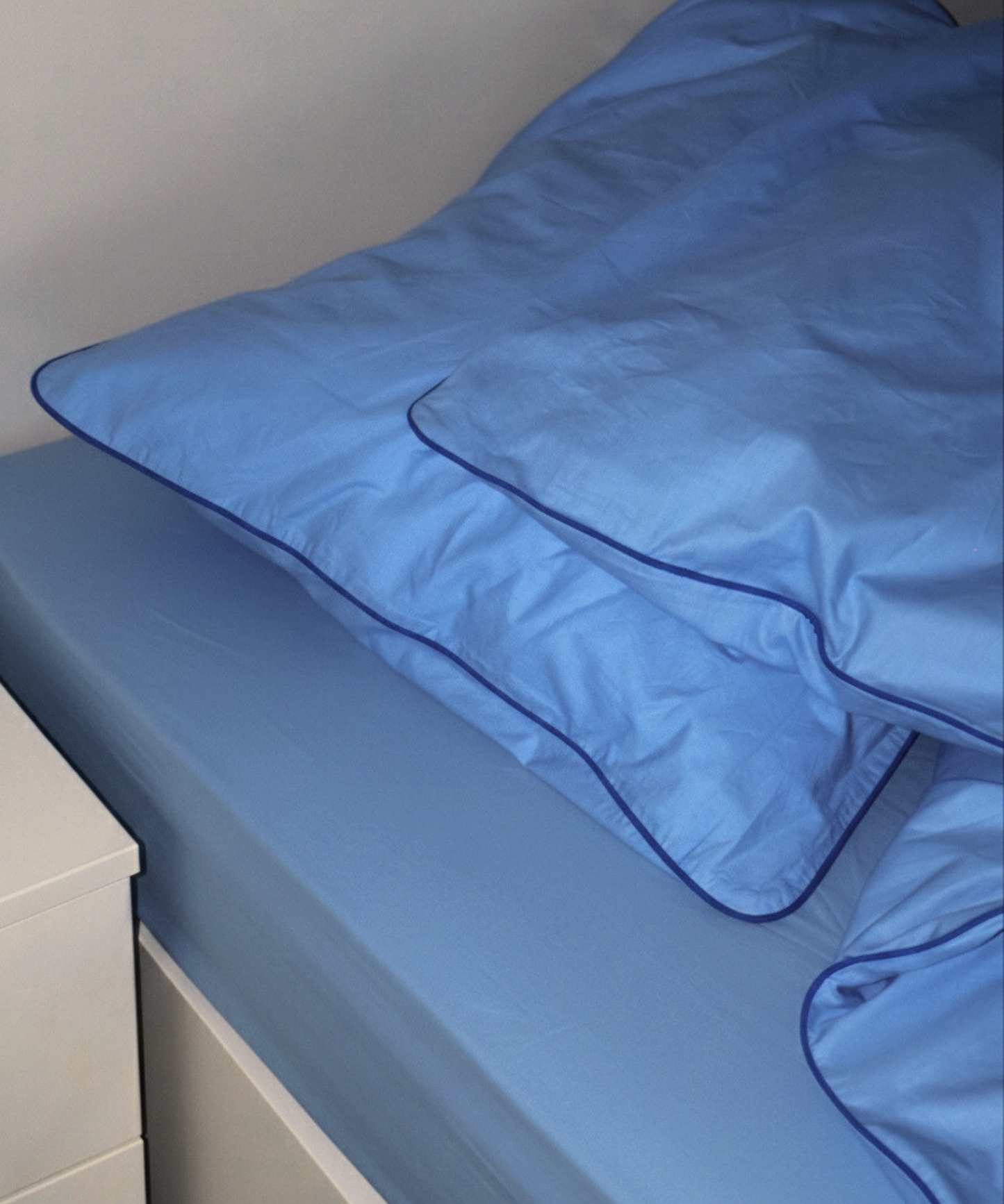 Blue bedding set including a pillowcase and duvet cover with dark blue piped edges, shown on a bed next to a white bedside table.