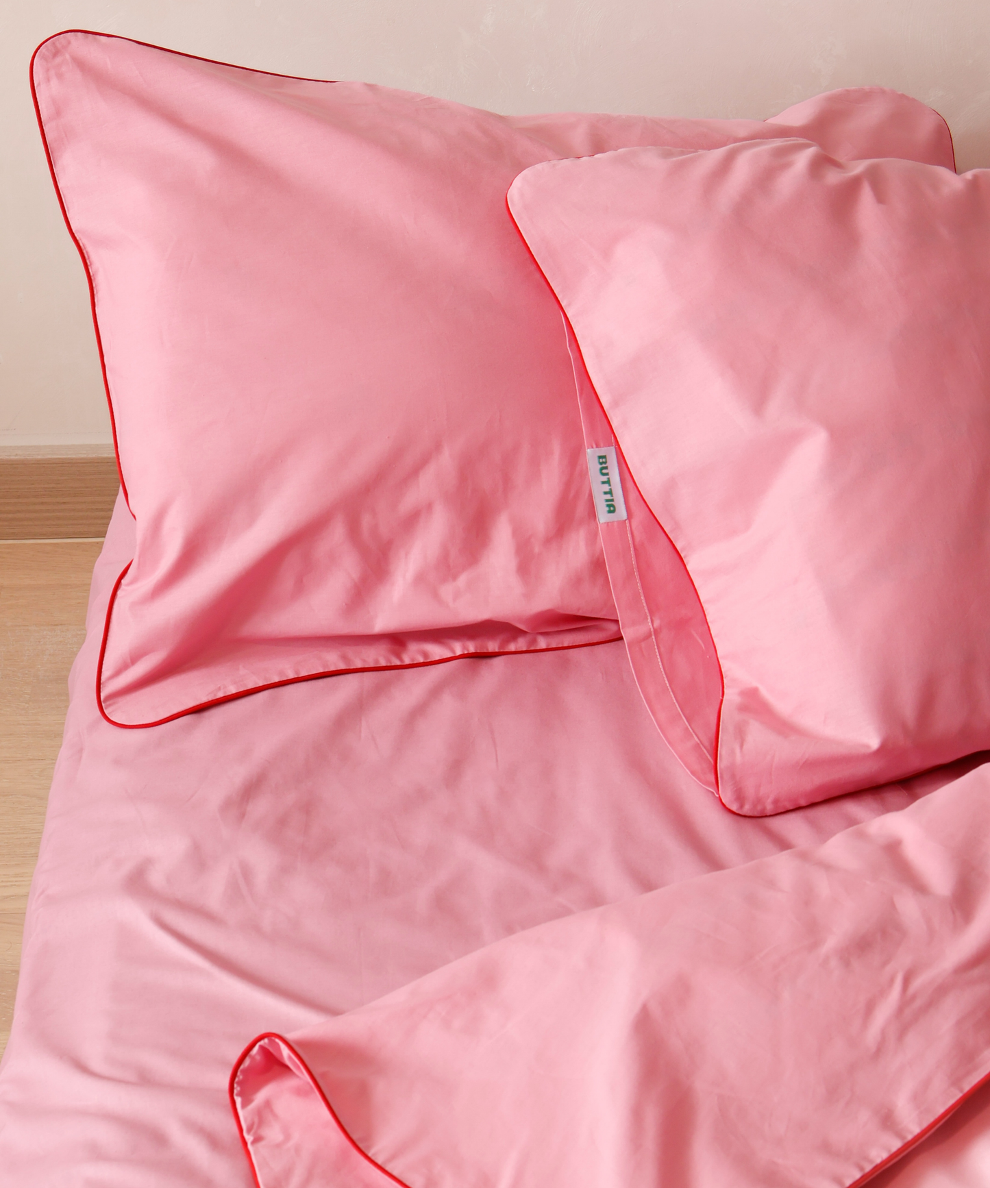Pink bed sheets and pillows with red piping on a wooden surface.