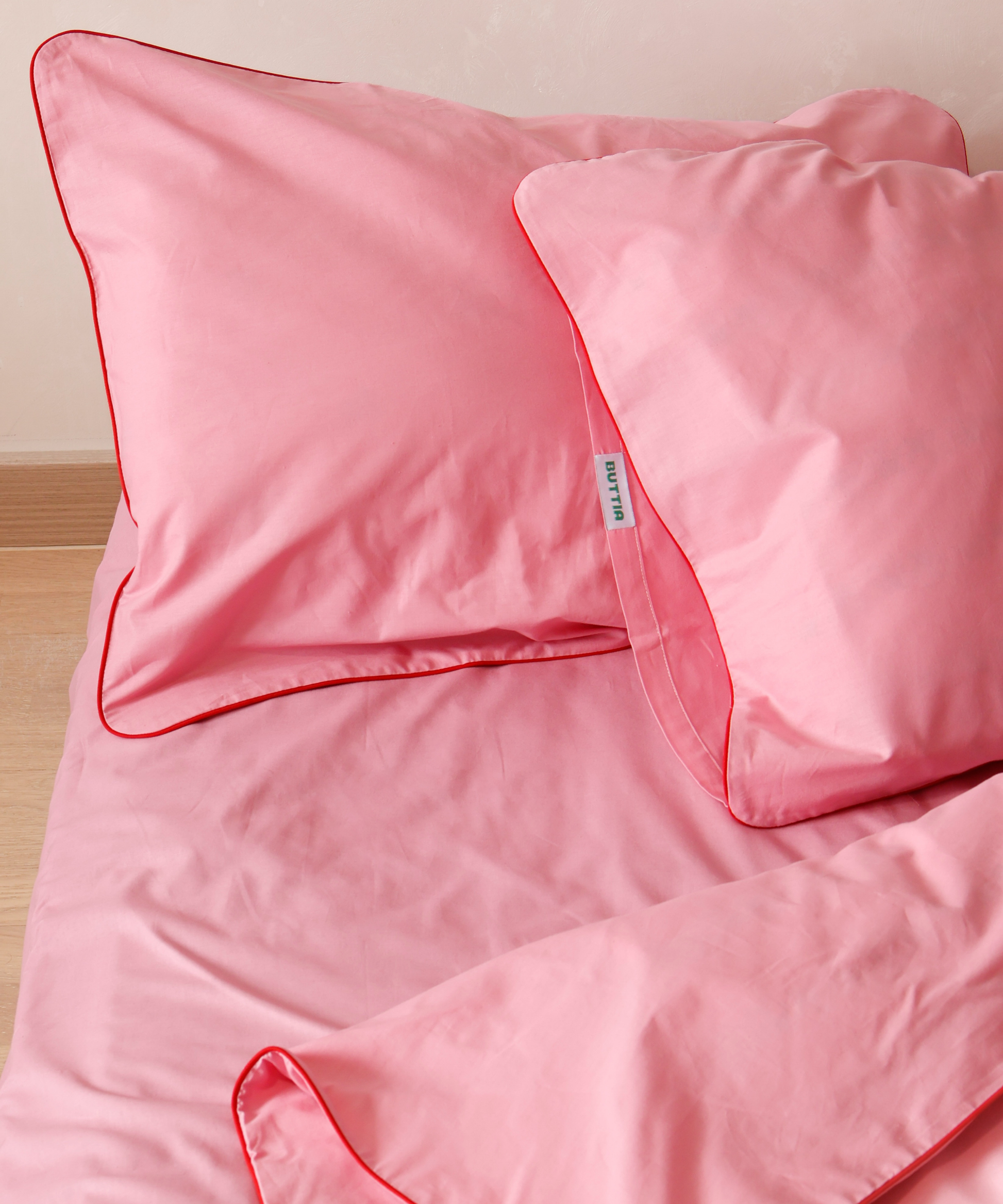 Pink bed sheets and pillows with red piping on a wooden surface.