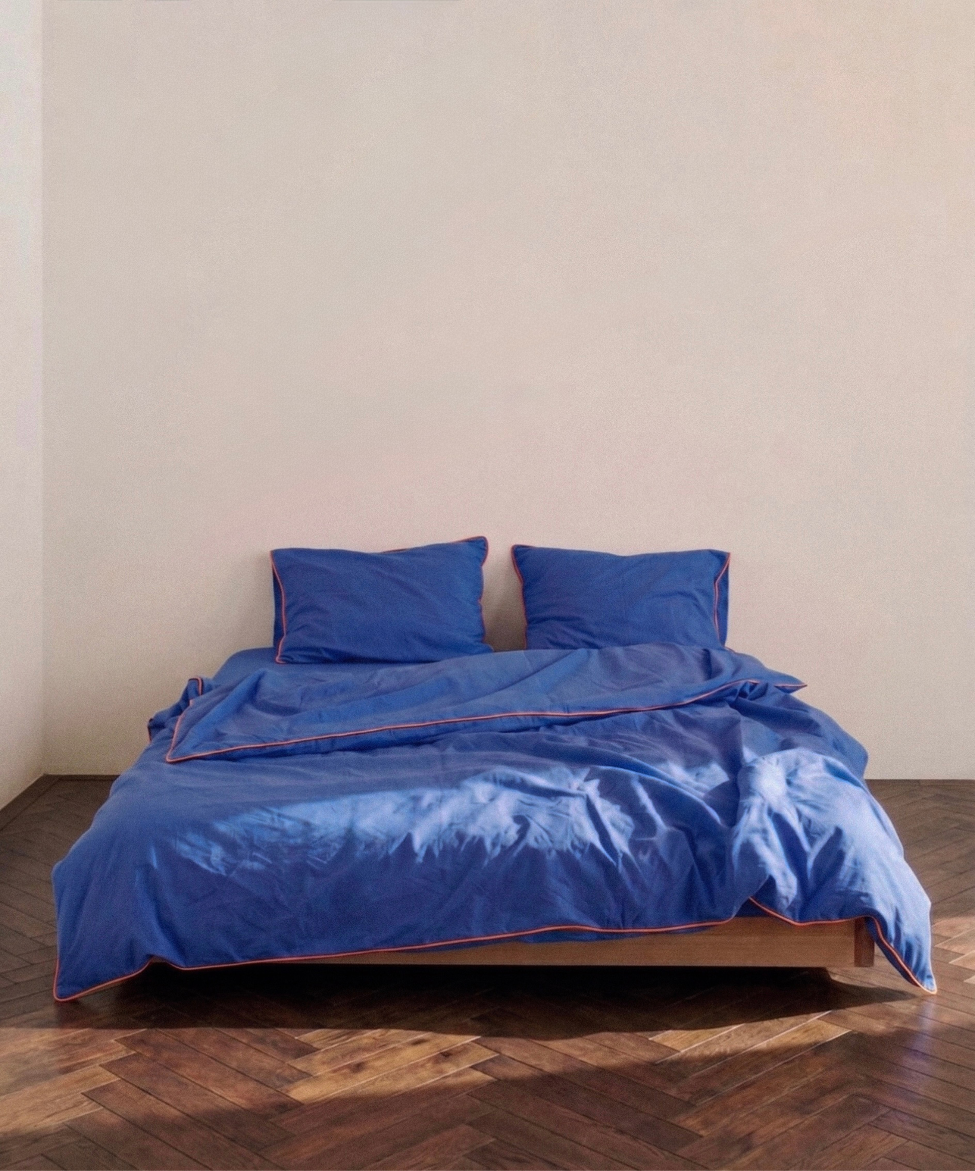 A bed dressed in a royal blue bedding set with matching pillows and a duvet, featuring a thin contrasting orange piping trim, set against a plain beige wall and dark wood parquet floor.