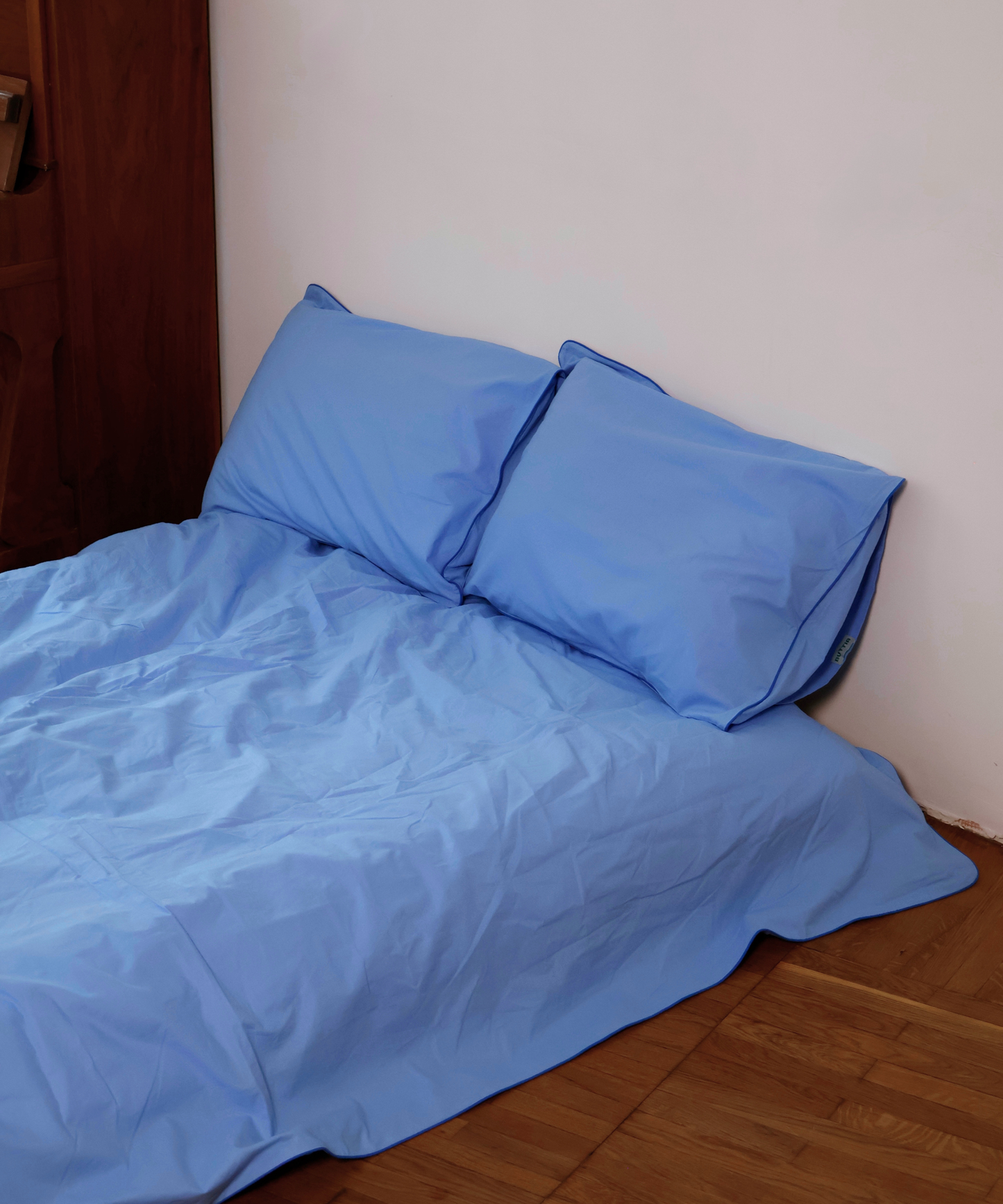 Light blue bedding set with two matching pillowcases on a bed in a bedroom corner with a white wall and wooden floor.