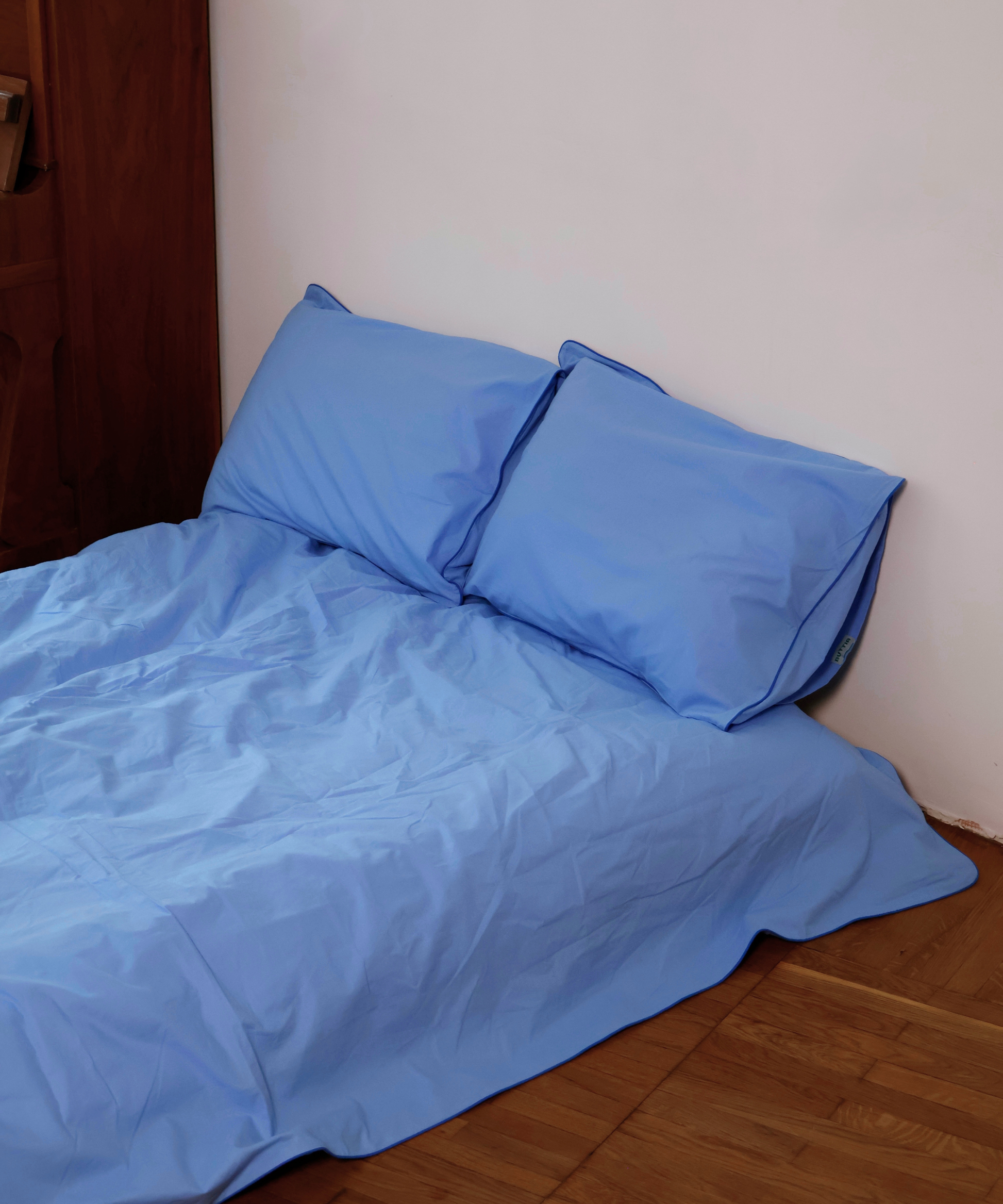 Light blue bedding set with two matching pillowcases on a bed in a bedroom corner with a white wall and wooden floor.