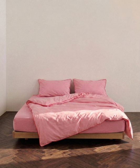 A bed dressed in a soft pink bedding set with two matching pillows and a rumpled duvet, set on a low wooden platform against a plain off-white wall and dark wood herringbone floor.