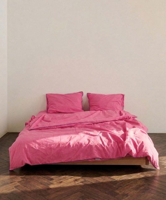 A bed dressed in bright pink bedding, including a duvet cover and two matching pillows, set against a plain off-white wall and dark wood parquet flooring.