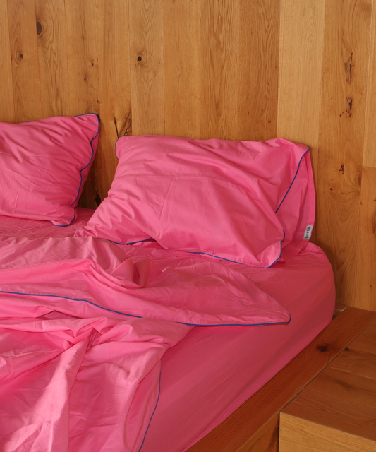 Bright pink bedding set with matching pillowcases and duvet cover, featuring thin blue piping trim, styled on a bed against a light wood-paneled wall.