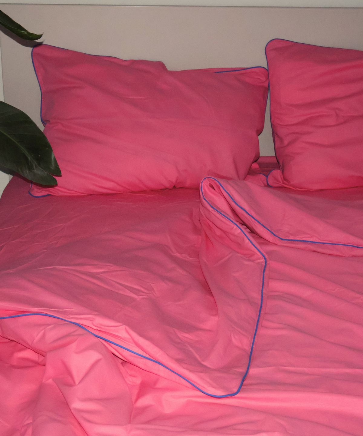 Pink bedding set with matching pillowcases and duvet cover, featuring a thin blue piping trim, shown on a bed with a neutral background.