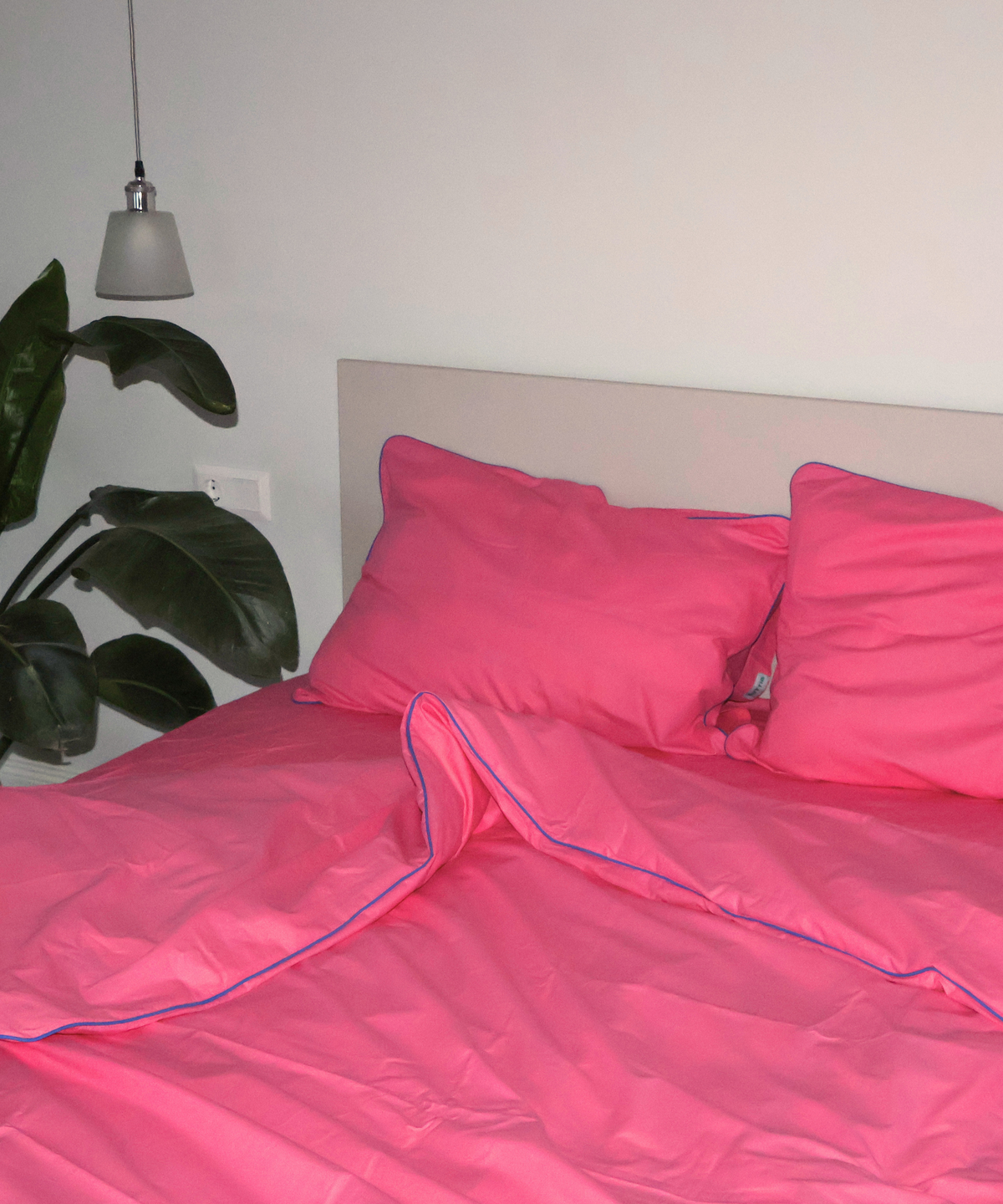 Bright pink bedding set with matching pillowcases and duvet cover featuring a thin blue piping trim, styled on a bed with a beige headboard in a bedroom setting.