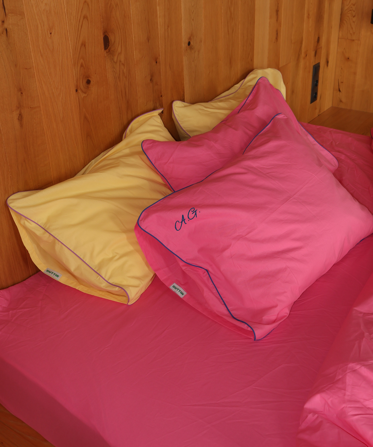 Bright pink and yellow bedding set on a bed with a wooden headboard, featuring two pink pillows with blue piping and one yellow pillow, with 'CAG' embroidered on the front pink pillow.
