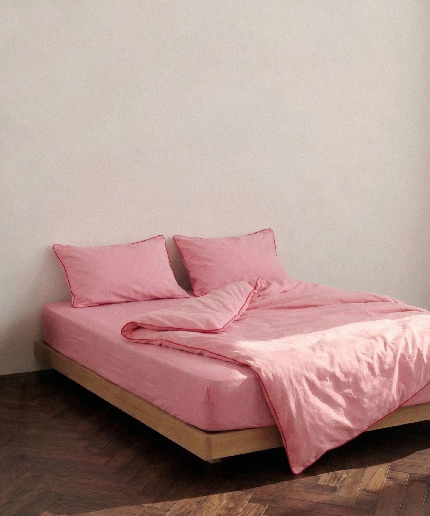 A bed dressed in a soft pink bedding set with matching pillows and duvet, set on a low wooden platform bed against a plain off-white wall and dark wood parquet floor.