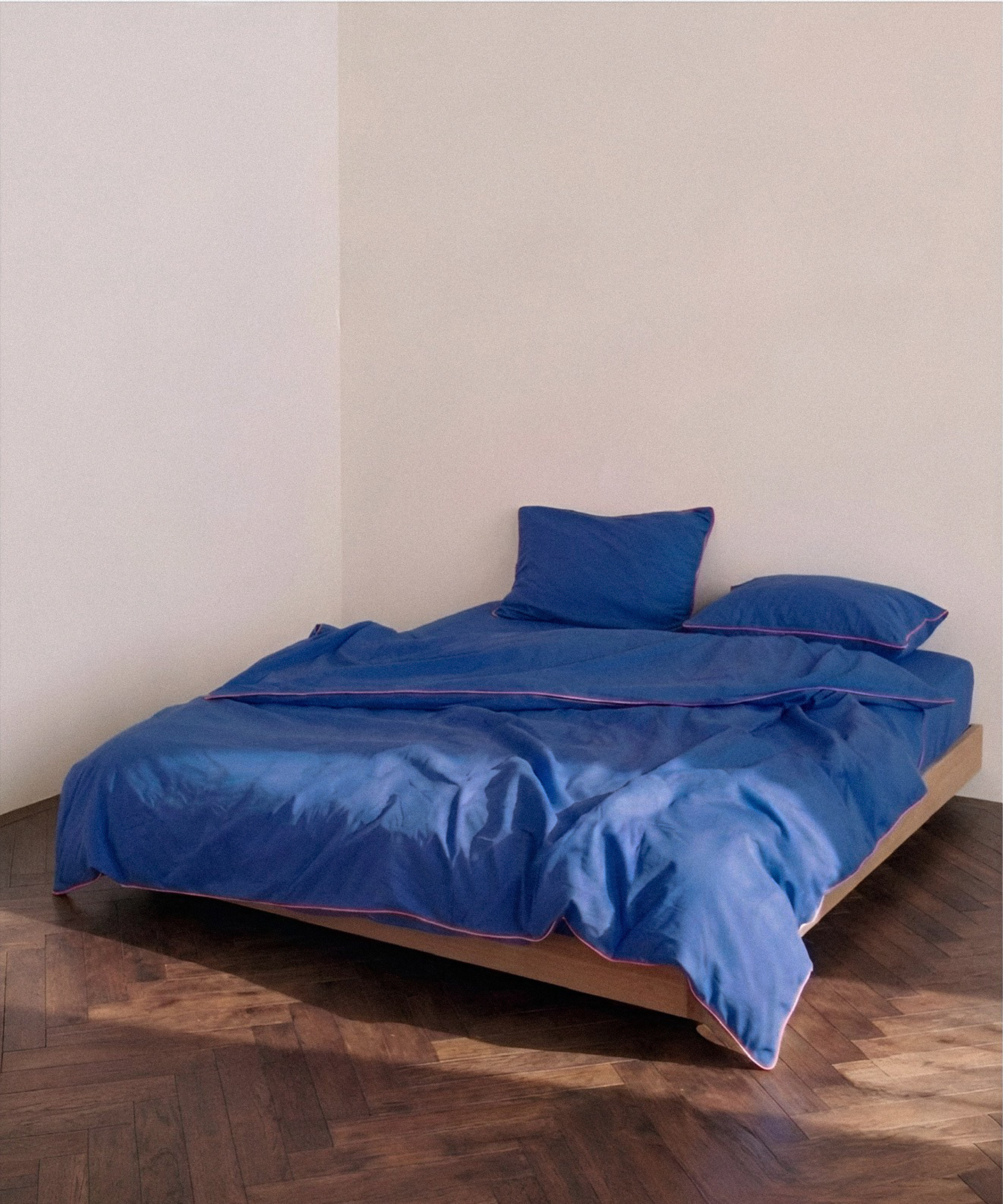 A bed dressed in a royal blue bedding set with matching pillows, set on a wooden platform bed frame in a minimalist room with beige walls and a dark wood parquet floor.