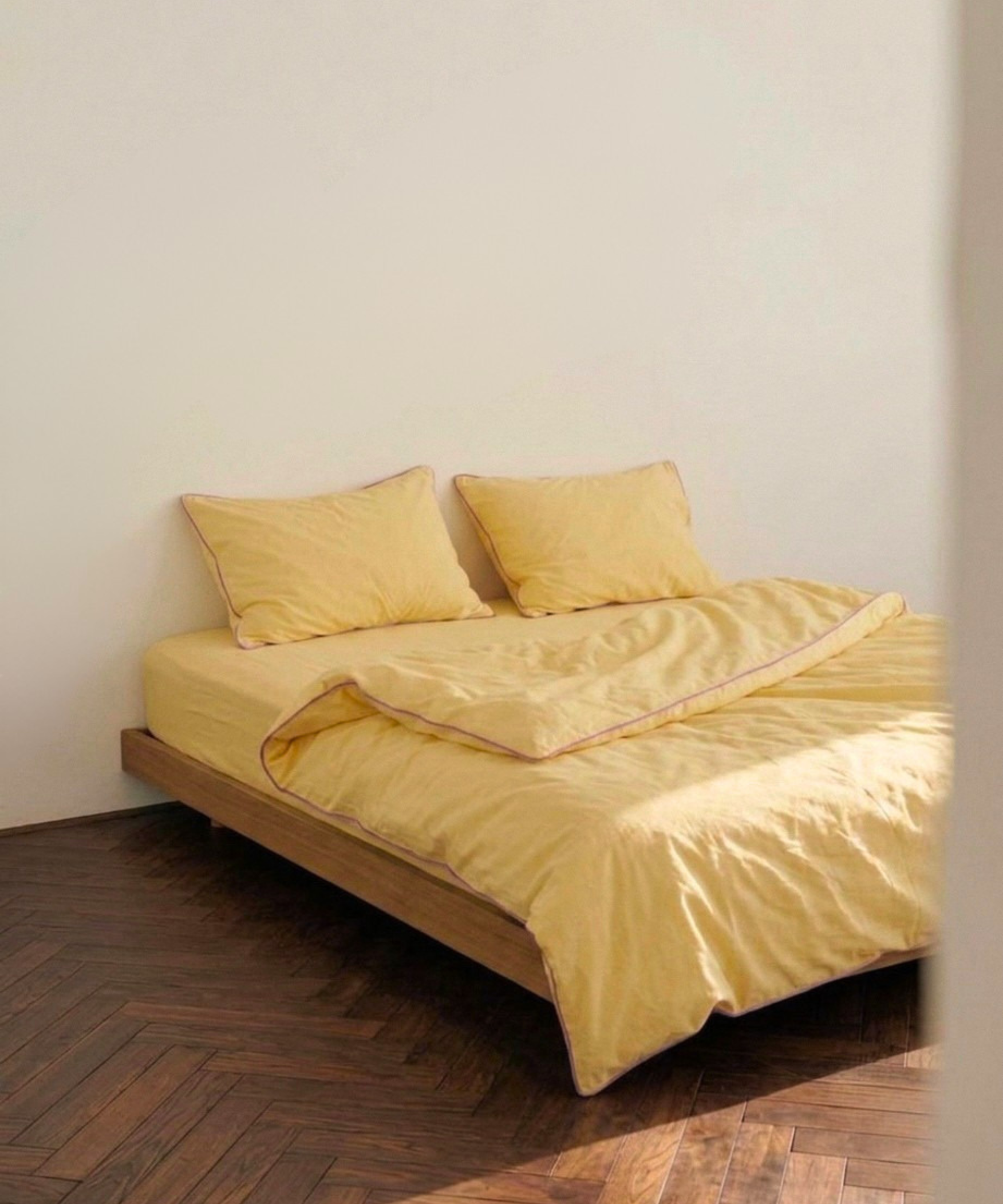 A bed dressed in soft yellow bedding, including a duvet cover and two matching pillows, set on a wooden platform bed frame against a plain off-white wall with a dark wood herringbone floor.