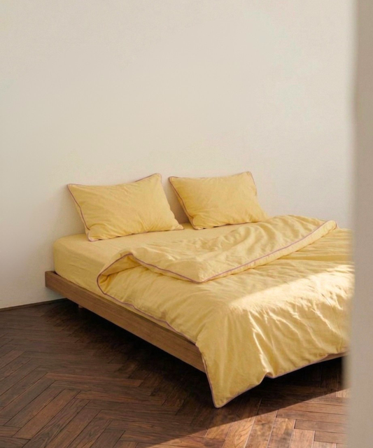 A bed dressed in soft yellow bedding, including a duvet cover and two matching pillows, set on a wooden platform bed frame against a plain off-white wall with a dark wood herringbone floor.