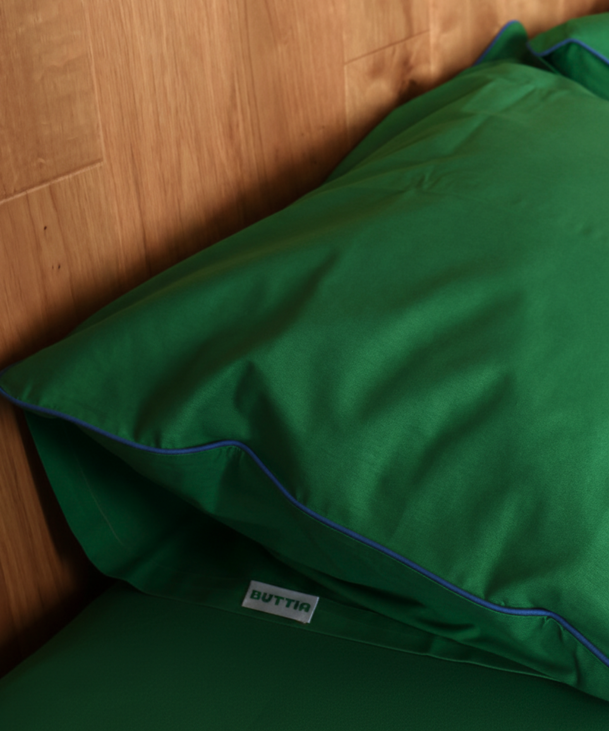 A close-up of a green pillowcase with blue piping trim, resting against a light wood headboard. A small white tag with the brand name 'BUTTIN' is visible near the bottom.