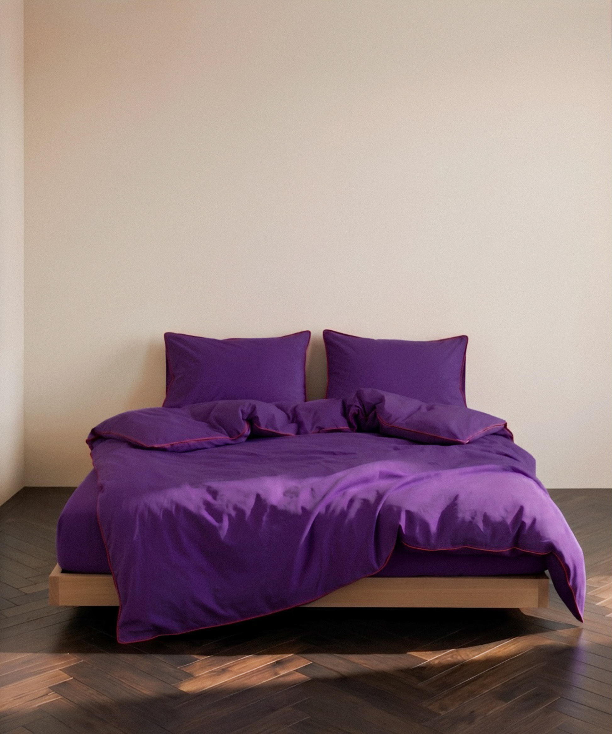 A bed dressed in a rich purple bedding set with matching pillows and duvet, set on a low wooden platform against a plain beige wall and dark wood parquet flooring.