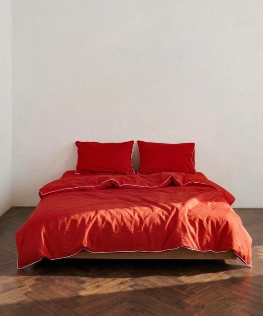 A bed dressed in a bright red bedding set with matching pillows and duvet, photographed in a minimalist room with a white wall and dark wood herringbone floor.