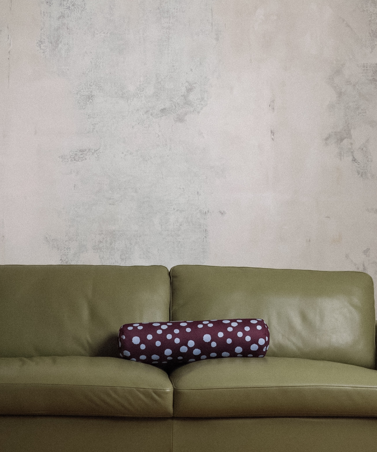 An olive green leather sofa against a textured plaster wall, featuring a burgundy cylindrical lumbar pillow with light blue polka dots.