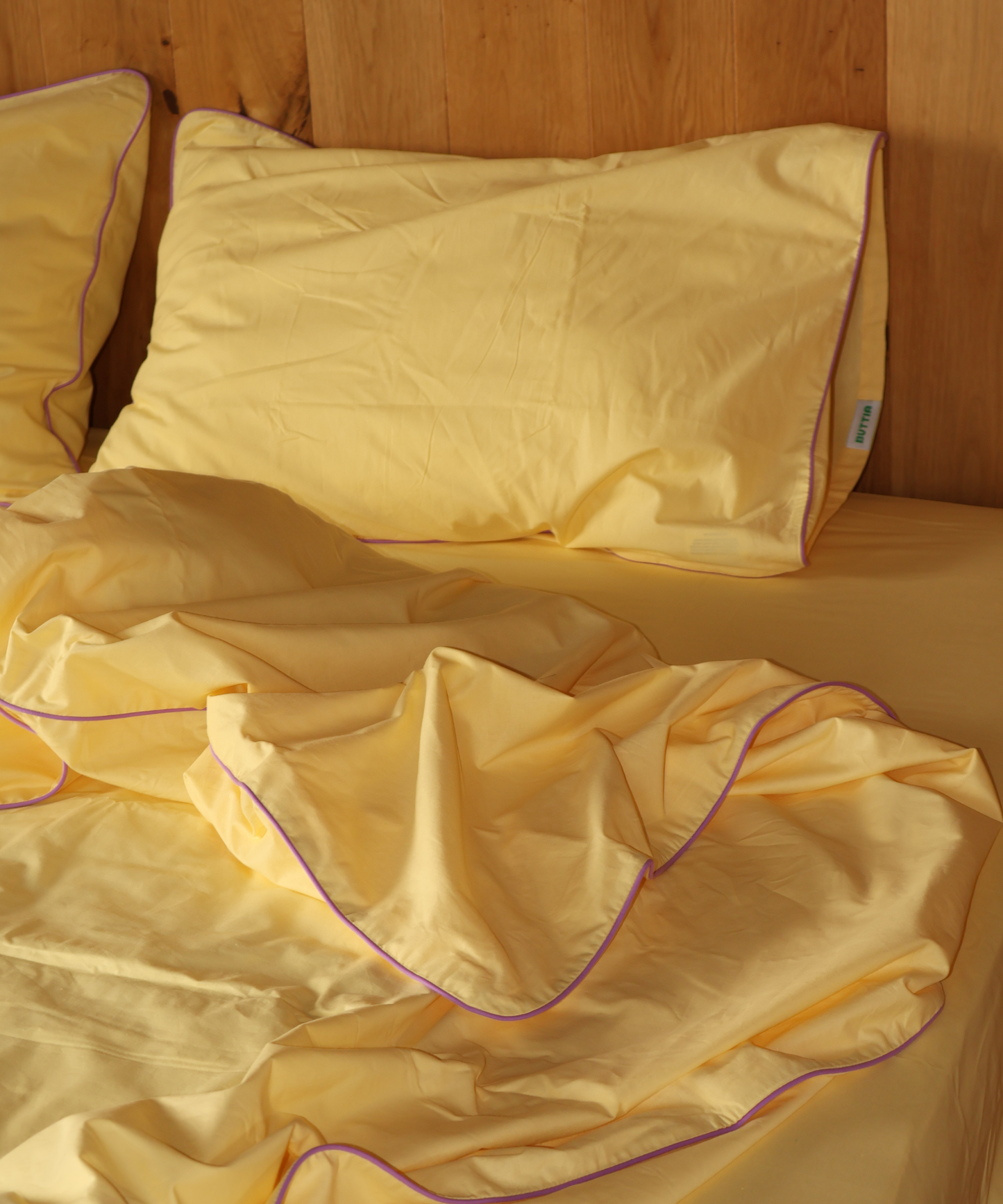 Yellow bedding set with a pillowcase and rumpled duvet cover on a bed against a wooden headboard, featuring a thin purple piping trim.