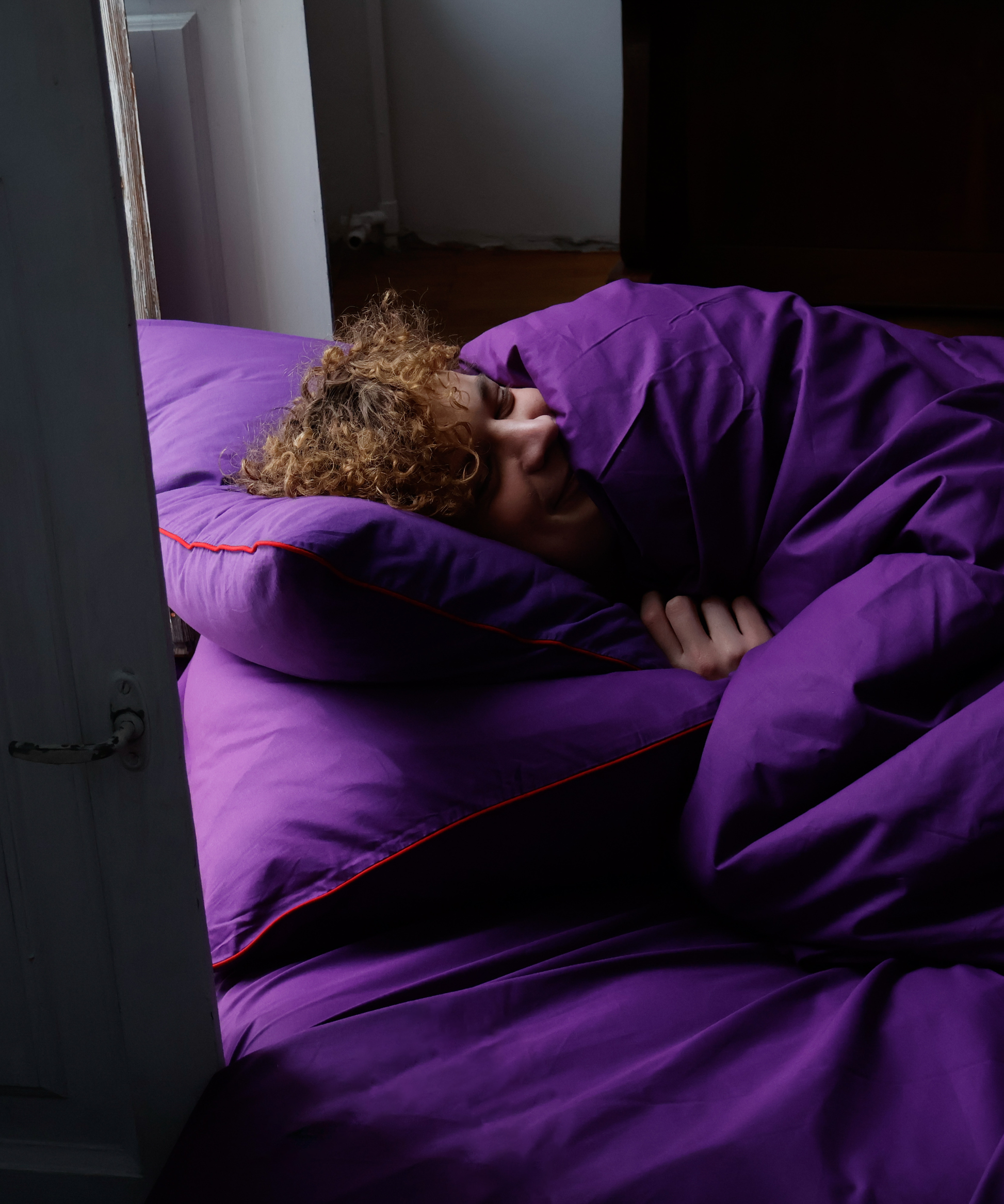 A person lying on a bed wrapped in a bright purple duvet and pillowcase with thin red piping trim, in a dimly lit bedroom.